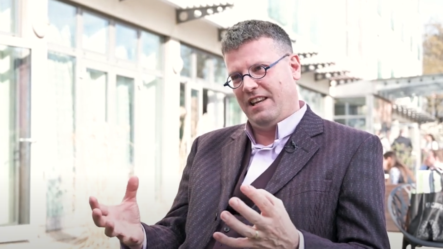 A person wearing glasses and a suit speaks animatedly while gesturing with both hands. They are seated outside in a sunlit area with a modern building in the background.