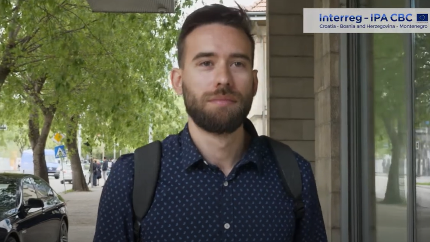 A man with a beard and wearing a blue patterned shirt carries a backpack while standing on a city sidewalk. Trees and parked cars are in the background. A sign for "Interreg - IPA CBC" is visible in the corner.