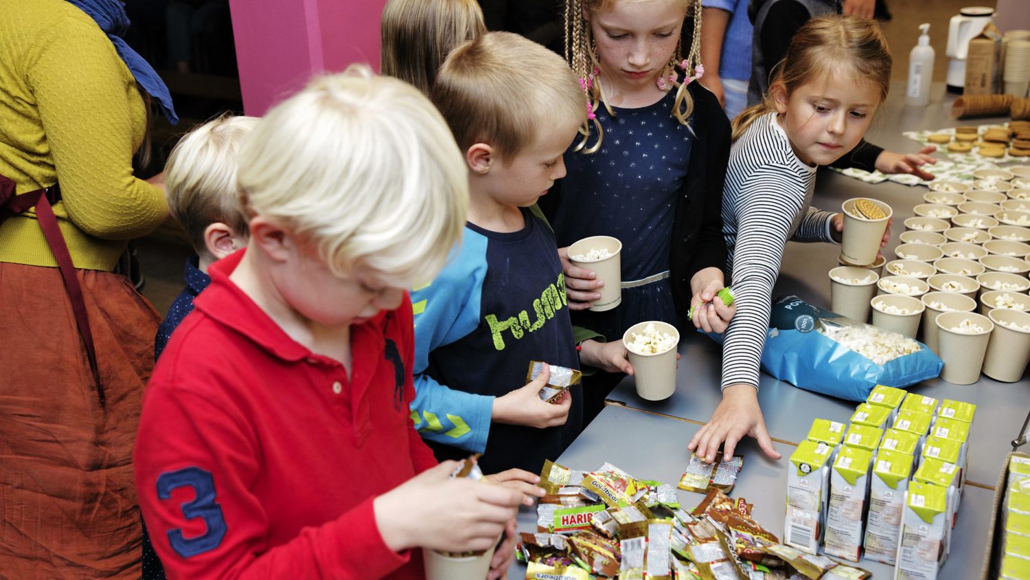 Children gather around a table selecting snacks and drinks, including juice boxes and candy. The atmosphere is lively, with bright colors and various items neatly arranged.