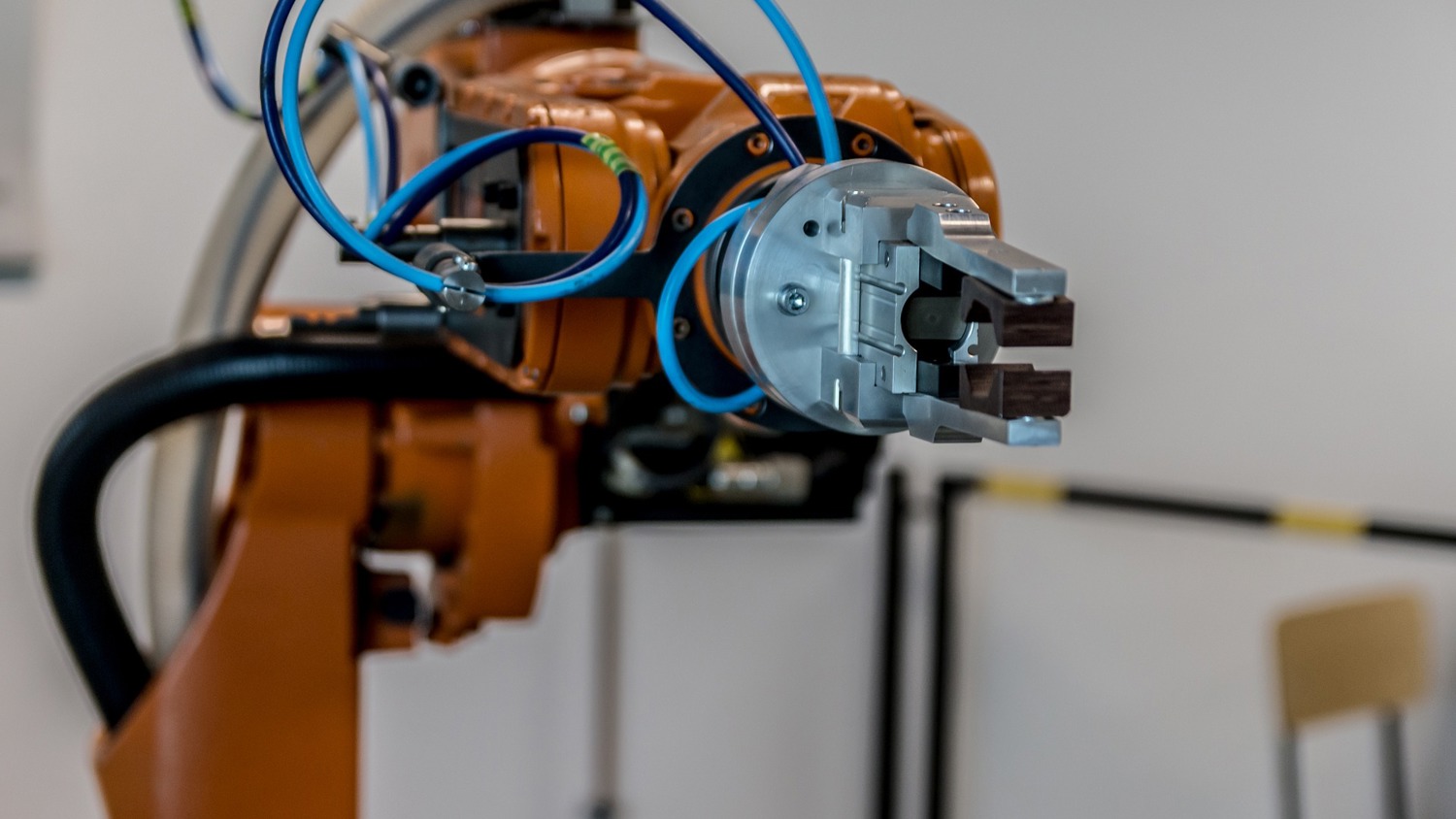 A close-up of an industrial robotic arm with a claw-like gripper. The arm is orange with blue cables attached, set in a blurred background, indicating a workshop or industrial setting.