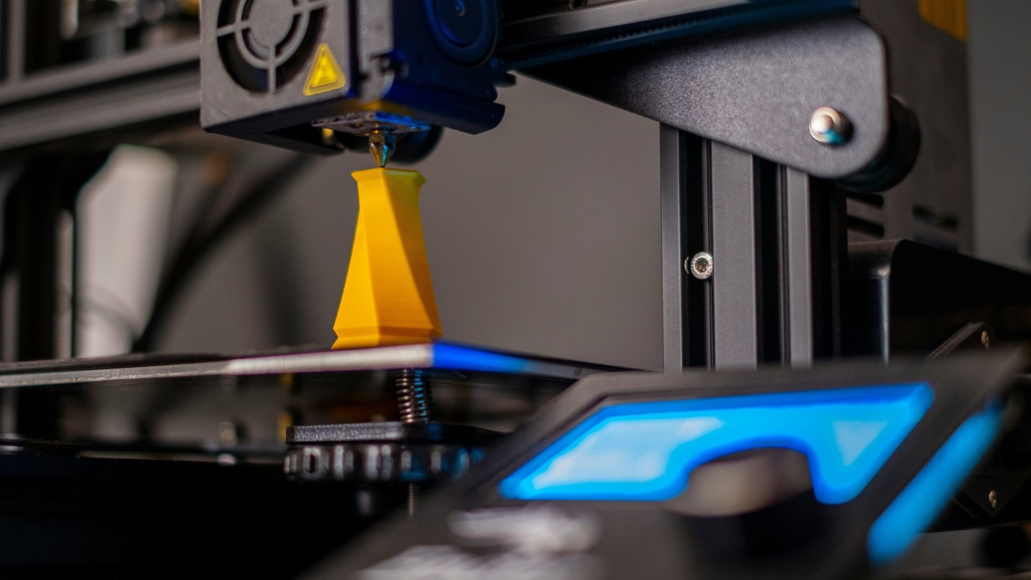 A close-up of a 3D printer in operation, creating a small yellow object. The machine's nozzle is visible above the object on the print bed. Part of the control panel with a lit-up screen is seen in the foreground.