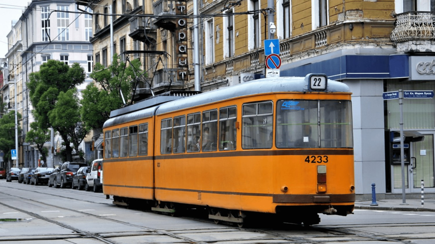 An orange tram marked "22" traveling down a street lined with historic buildings. Cars are parked along the road, and tramlines are visible on the pavement. A blue sign with a white arrow points upward.