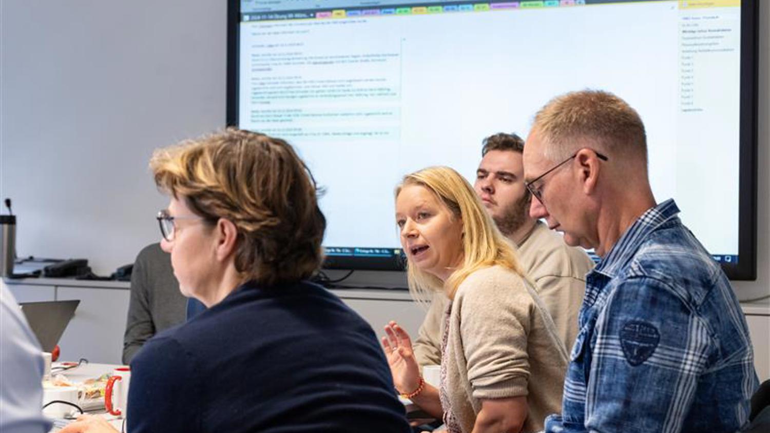 A group of people sit at a conference table engaged in discussion. A woman in the center talks while others listen. A large screen displays a document in the background. Coffee mugs and laptops are on the table.