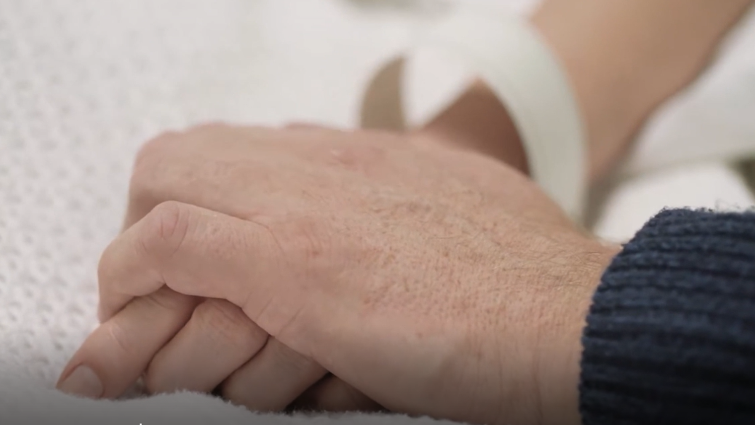 A close-up of two hands clasped together. One hand wears a hospital wristband and rests on a white textured blanket, while the other hand appears to offer comfort, wearing a dark blue knitted sleeve.