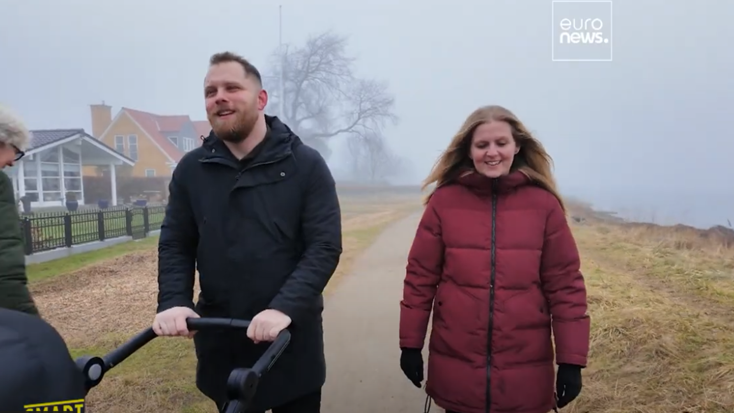 A man pushes a stroller beside a woman on a foggy path. Another person walks nearby. They are all dressed warmly. Houses and leafless trees are visible in the background. The Euro News logo is in the top-right corner.