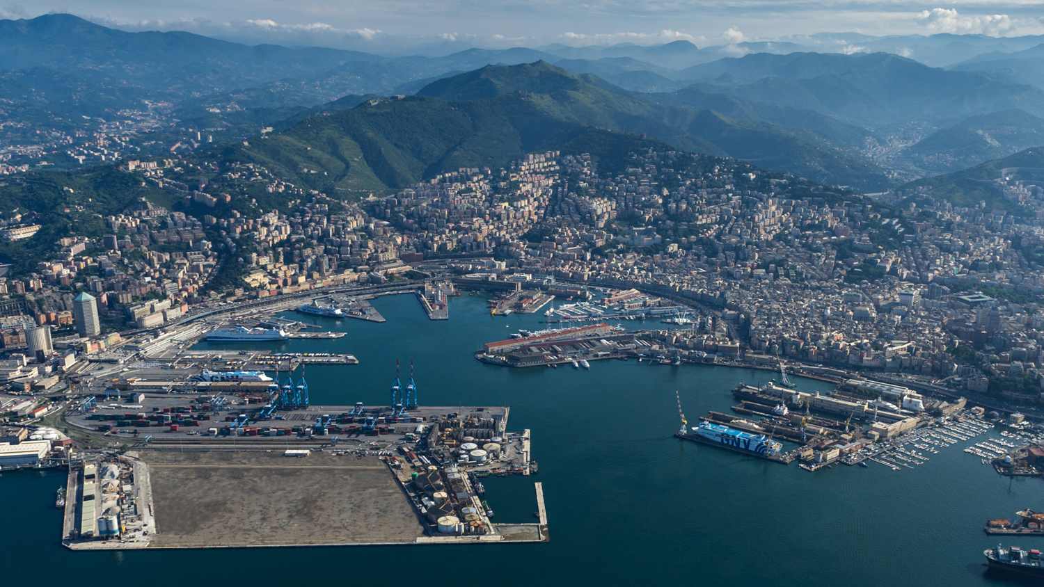 Aerial view of a coastal city with a large port. The area shows several docks, cranes, and ships. Surrounding hills and mountains form the backdrop, with urban development expanding towards them. The water is calm, and the sky is partly cloudy.