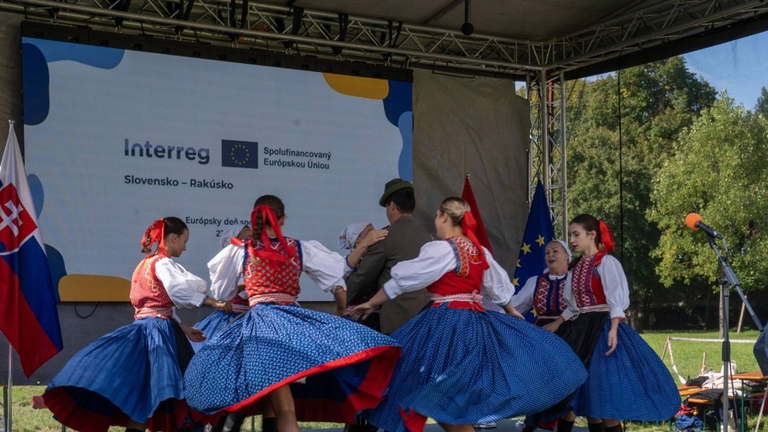 A group of people in traditional Slovak folk costumes dance on an outdoor stage. Behind them is a screen with Slovak and EU flags and text about a Slovakia-Austria EU-funded program. Trees and blue sky are in the background.