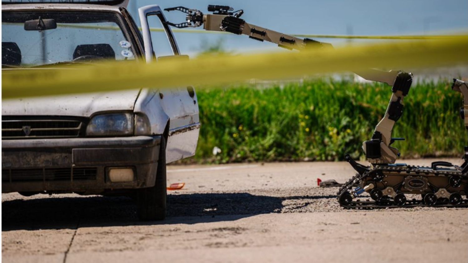 A remote-controlled robotic arm approaches a parked, weathered car behind yellow caution tape on a sunny day, set against a backdrop of greenery.