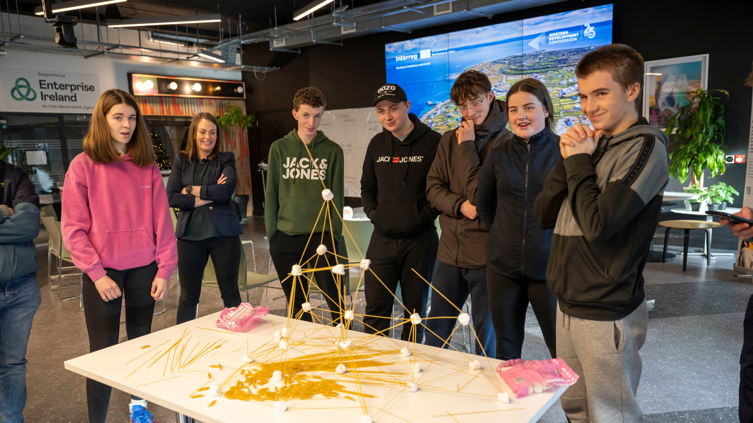 A group of young people stand around a table, observing a tall structure made of spaghetti and marshmallows. They appear engaged and amused. The setting looks like a modern indoor workspace.