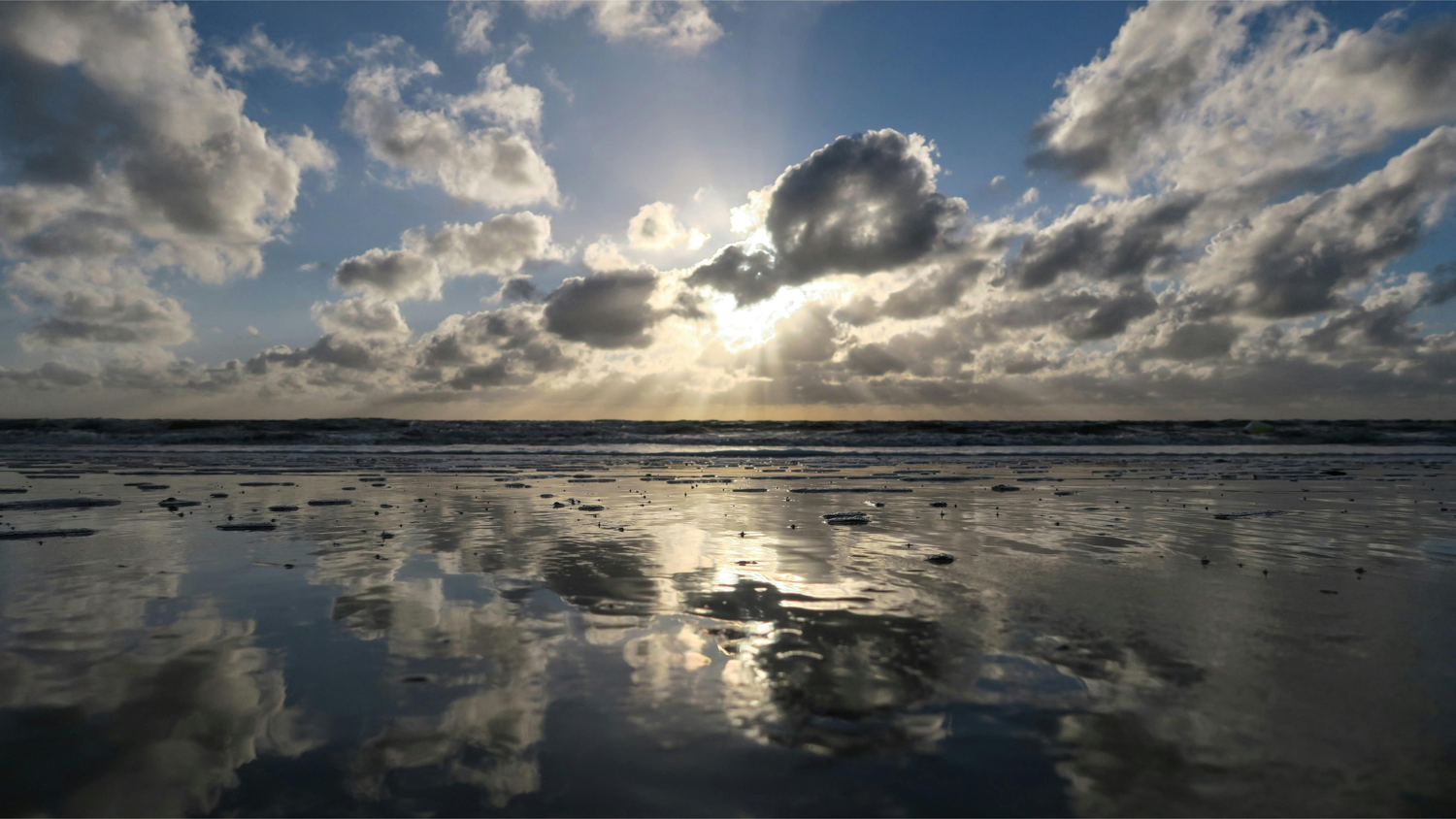 Sun setting over a calm sea; clouds partly cover the sun, casting reflections on the wet sandy shore.