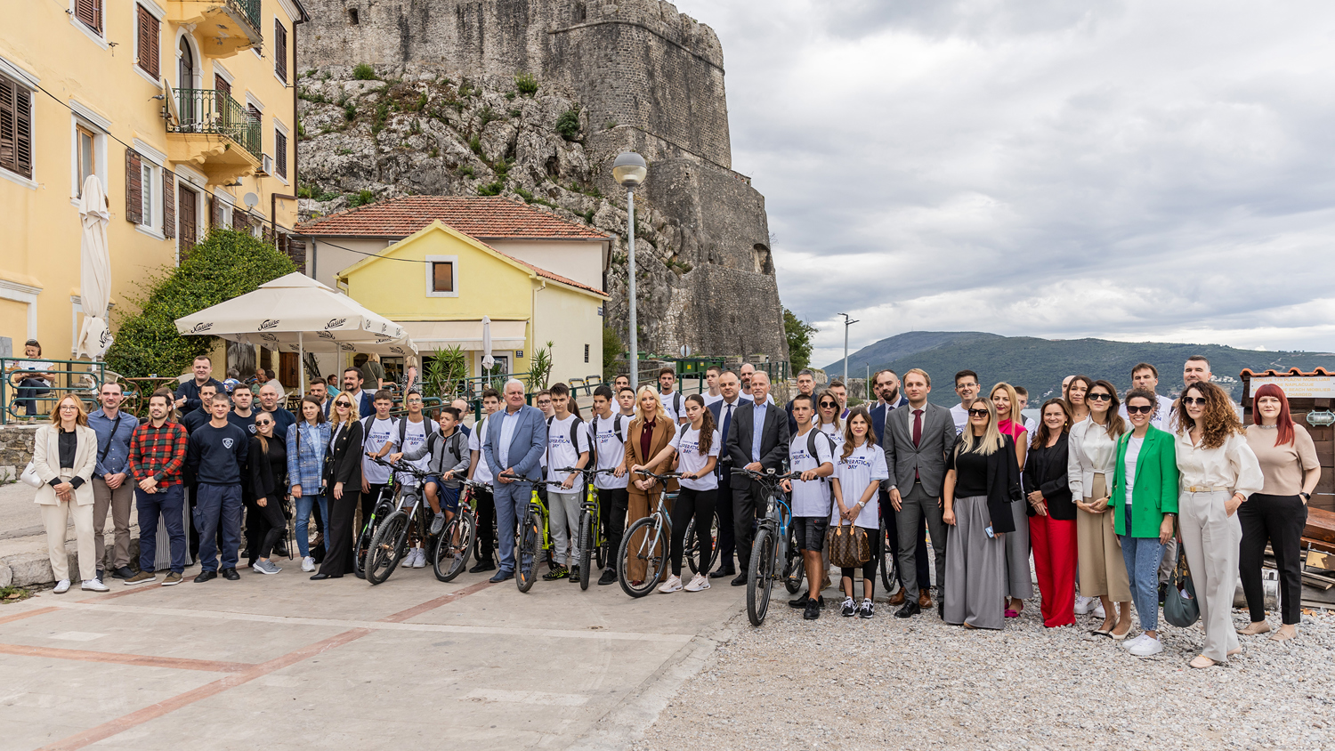 A large group of people, some with bicycles, stand together in front of a historic stone building on a cloudy day. The scene includes a mix of men and women, wearing casual and formal attire, with a rocky hill in the background.