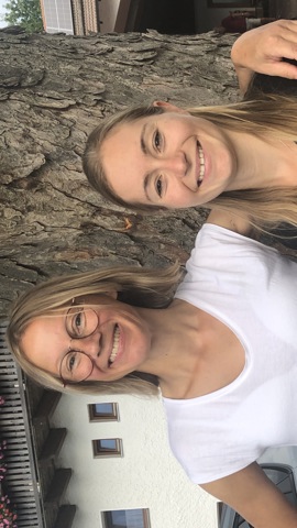 Two women stand smiling in front of a tree. The woman on the left wears glasses, a white t-shirt, and gray jeans; the woman on the right wears a black t-shirt and red pants. They are outside next to a building.