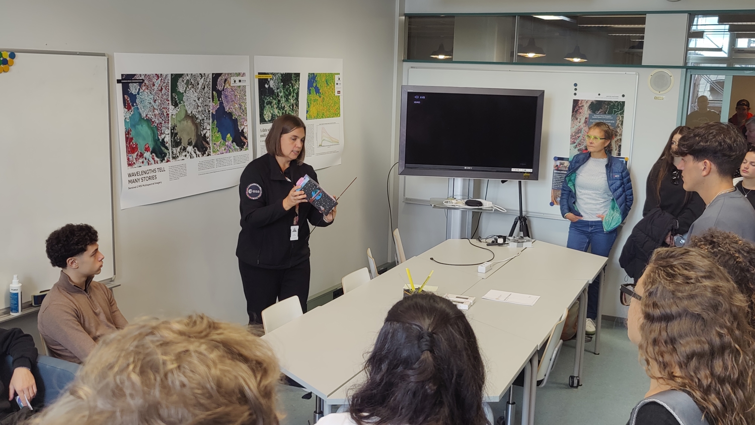 A group of people stand around a conference table in a meeting room. A woman is holding a book and speaking to the group. There are large images on the walls and a flat-screen TV is mounted behind her.