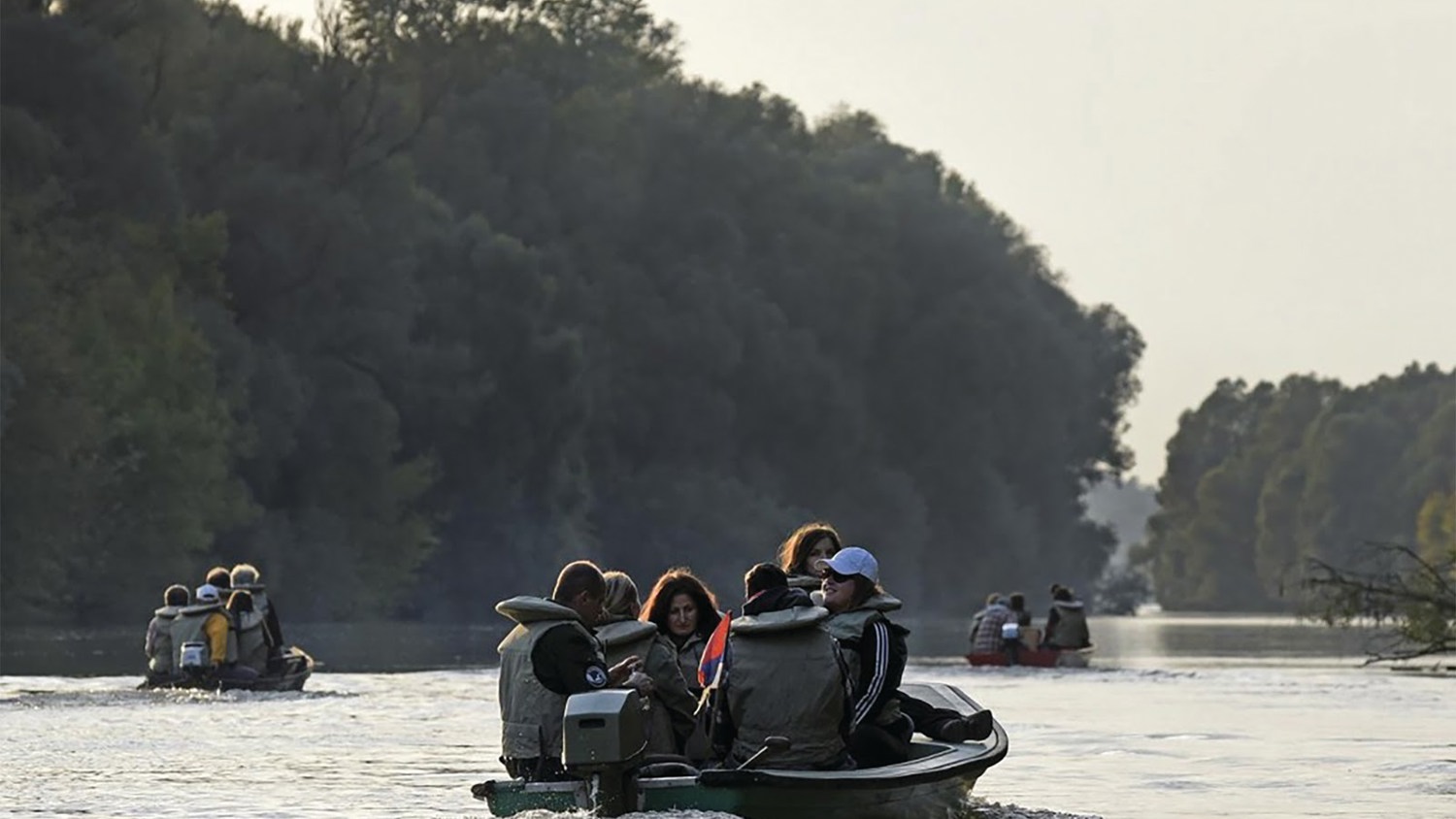 A group of people traveling in small boats on a wide river, surrounded by lush forests. The scene is serene with trees lining the riverbanks, creating a peaceful, natural atmosphere. The sky is overcast, adding to the tranquility.