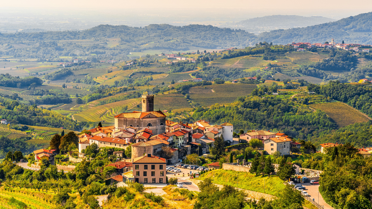 A scenic view of a quaint village nestled among rolling hills and vineyards. The village features a prominent church with a tall tower and red-roofed buildings. In the background, lush green hills extend towards the horizon under a hazy sky.