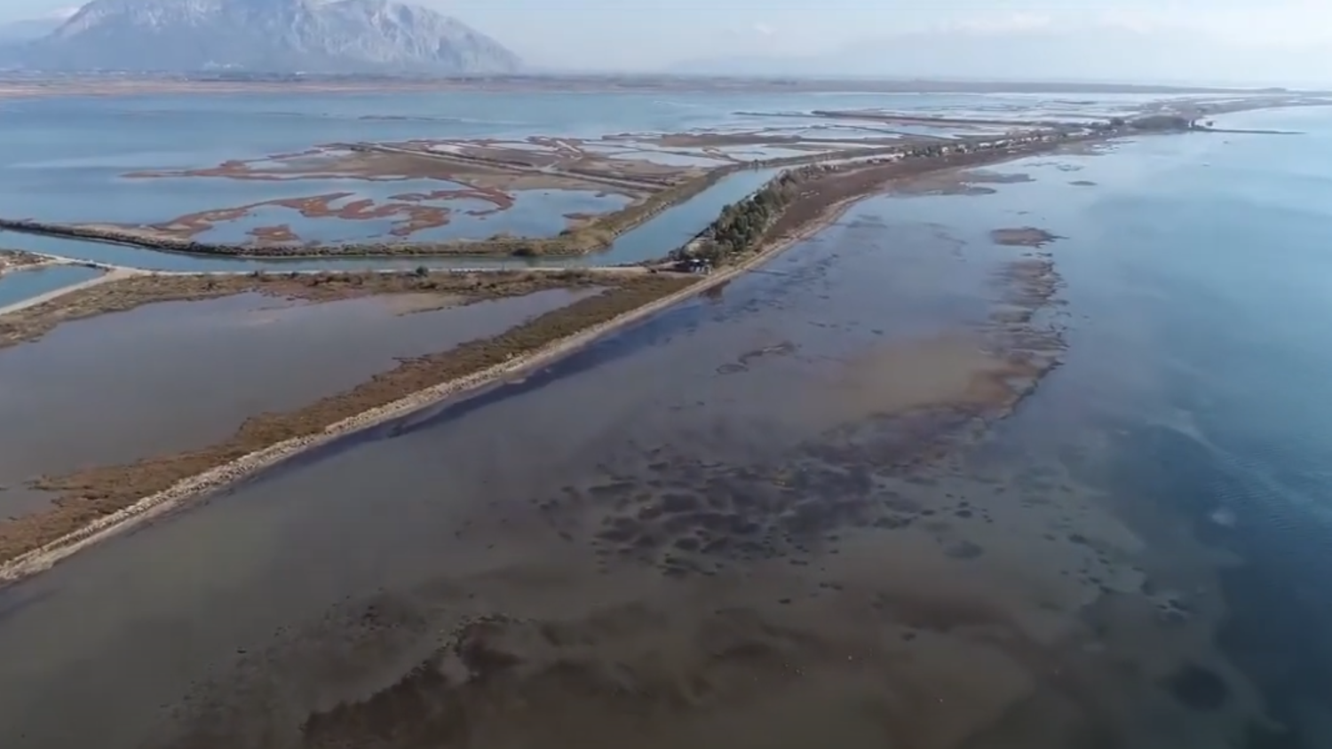Aerial view of a narrow strip of land surrounded by water, with salt marshes and a distant mountain under a clear sky. The landscape features a blend of water channels and patches of vegetation.