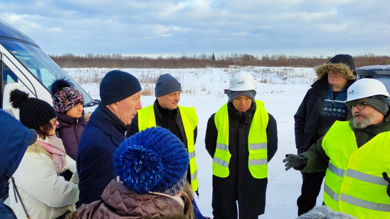 A group of people wearing winter clothes and high-visibility vests stand in a snowy outdoor setting. Two individuals are wearing white hard hats, and one is gesturing while speaking to the group. A van is visible on the left.