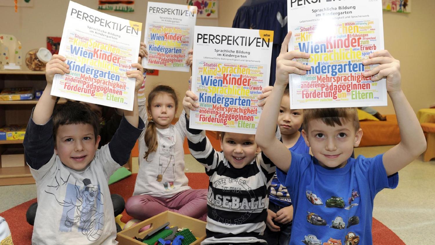Children sitting on the floor hold up colorful magazines titled "Perspektiven" in a classroom setting. They are smiling, and a box of toys is in front of them, suggesting a playful learning environment.