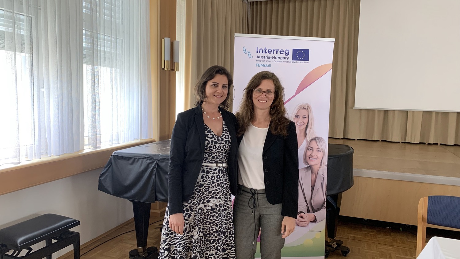Two women standing and smiling in front of a banner with text and images. The woman on the left is wearing a black blazer and polka dot dress, while the woman on the right is in a black jacket and gray pants. They're in a room with a piano and curtains.