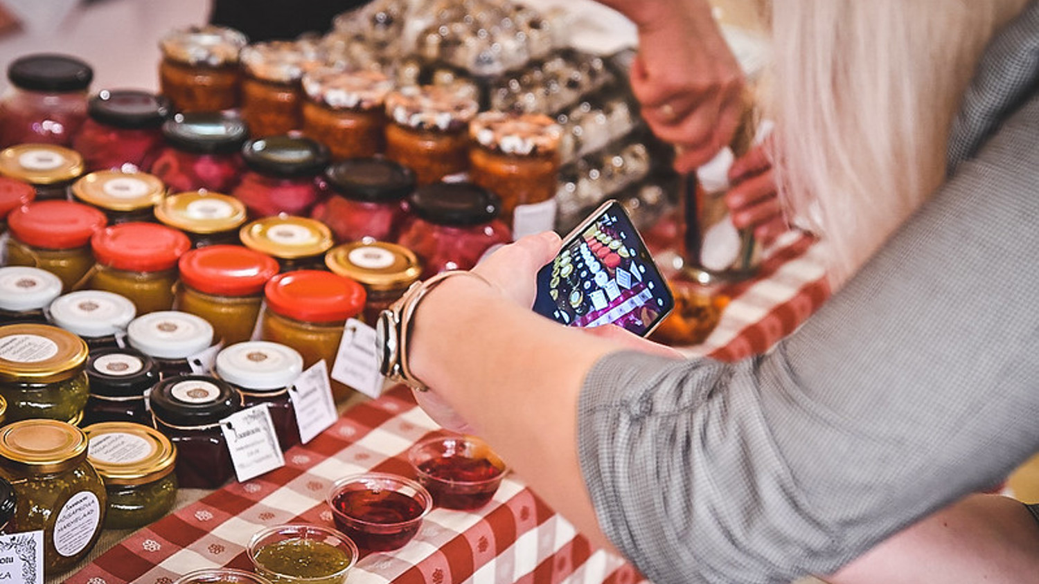 Person taking a photo of various colorful jars on a plaid tablecloth at a market stall. Other containers of samples are in the foreground, and another person is handling a jar behind the table.
