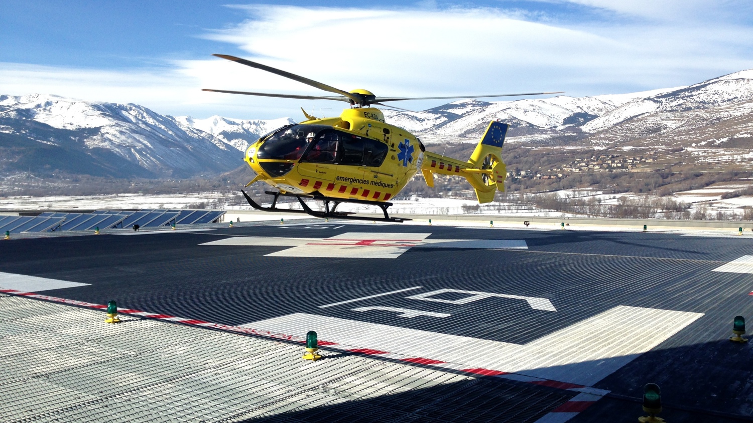 A yellow medical helicopter with a blue star of life symbol is taking off from a helipad in a mountainous area. The landscape features snow-capped mountains and a clear blue sky in the background.