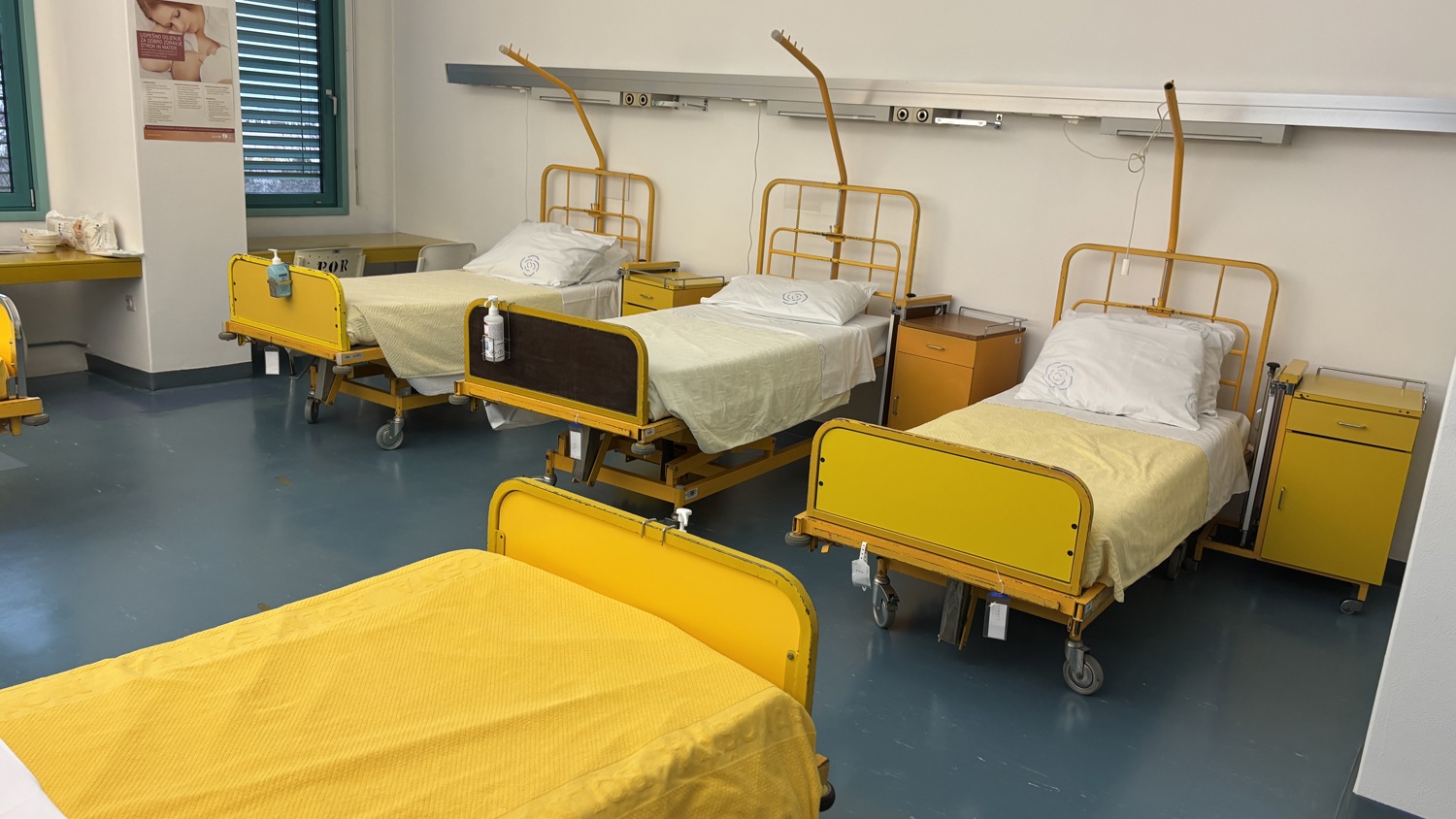 Four hospital beds with yellow blankets are arranged in a room. Each bed has a white pillow. Medical equipment is mounted on the wall behind the beds. A window with blinds is on the left. The floor is a bluish shade.