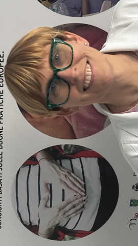 A person with short hair and glasses, wearing a white coat, smiles in front of a poster written in Italian and Slovenian. The poster describes a health project related to physiological pregnancy.
