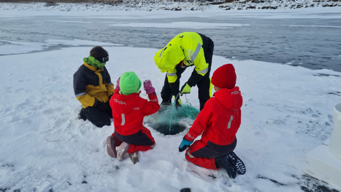 People in winter clothing gather around a hole in the ice, engaged in ice fishing. Behind them is a frozen lake. Snow covers the ground and the landscape is wintry. The group includes adults and children wearing bright jackets.