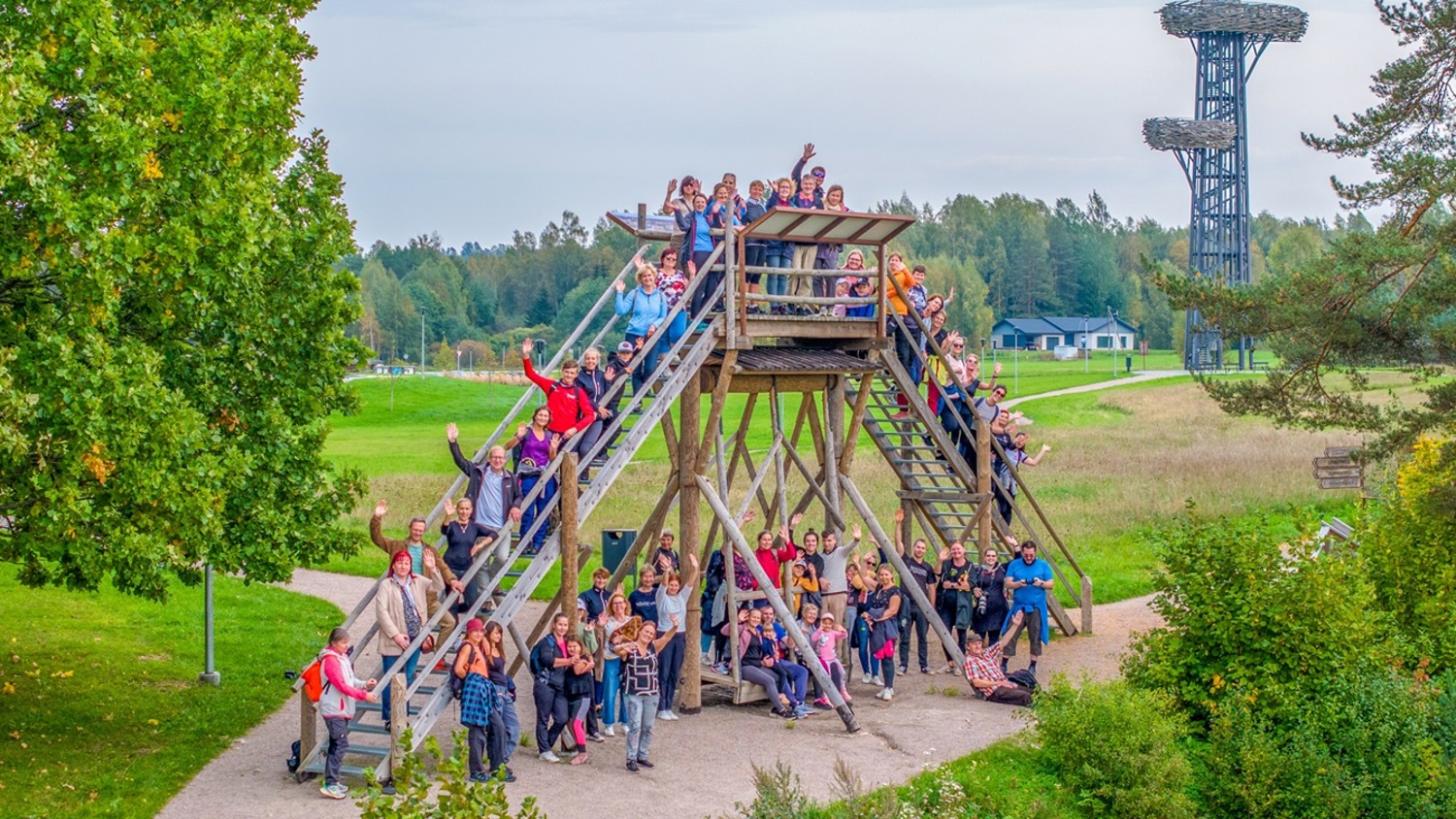 A large group of people pose cheerfully on a tall wooden structure with stairs, surrounded by green trees and grass. In the background, a tall metal observation tower is visible under a cloudy sky.