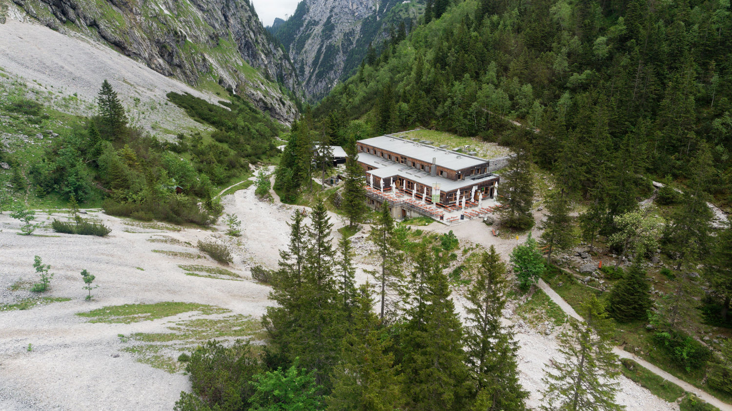 A large, modern building with a flat roof is nestled among lush green trees in a mountain valley. Surrounding rocky and forested slopes rise toward a cloudy sky, creating a serene, secluded setting.