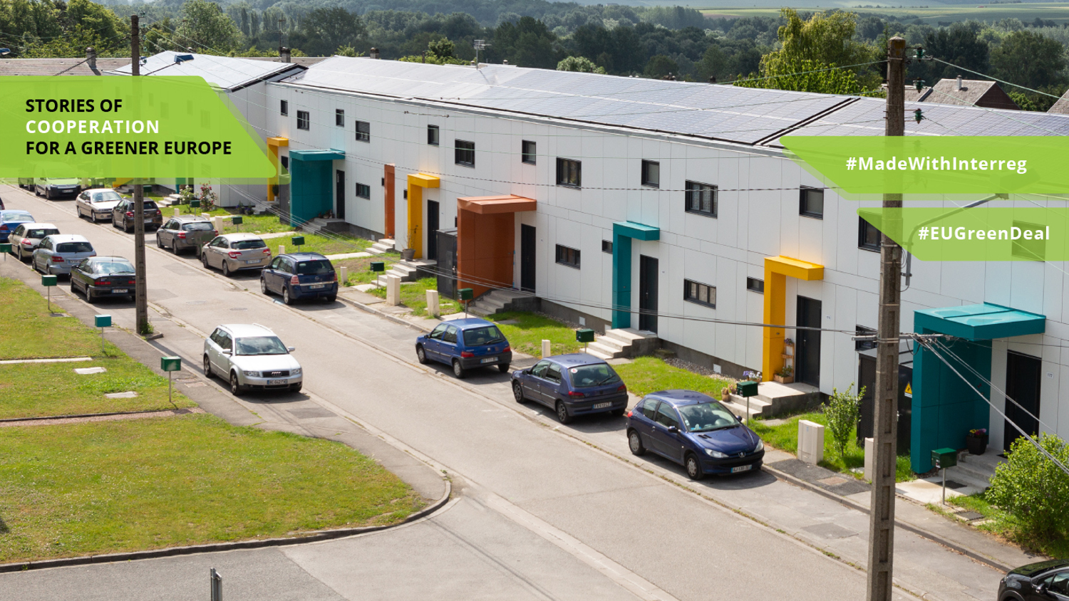 A row of modern houses with colorful doors and solar panels on the roofs, set along a quiet street. Green banners with text read "Stories of Cooperation for a Greener Europe" and hashtags #MadeWithInterreg and #EUGreenDeal are visible.
