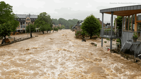 A flooded street with fast-moving brown water flows between houses and trees. On the right, a building with a balcony overlooks the scene. The sky is overcast, and debris is visible in the water.