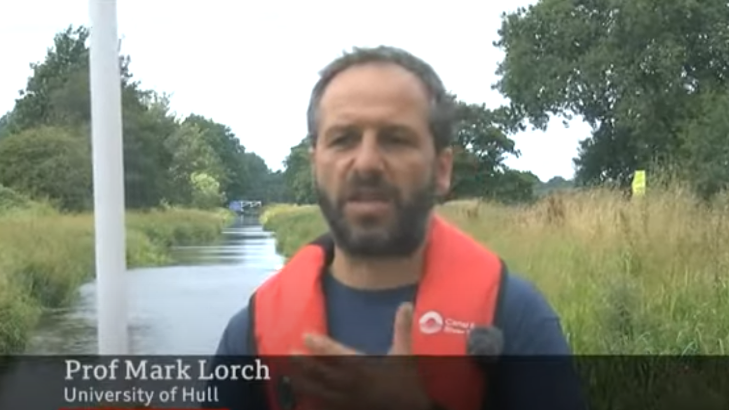 A person wearing a red life jacket stands by a river, surrounded by grass and trees. Text on the image says "Prof Mark Lorch, University of Hull.