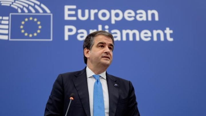 A man in a suit stands at a podium with a microphone, in front of a blue background displaying the European Parliament logo and text.