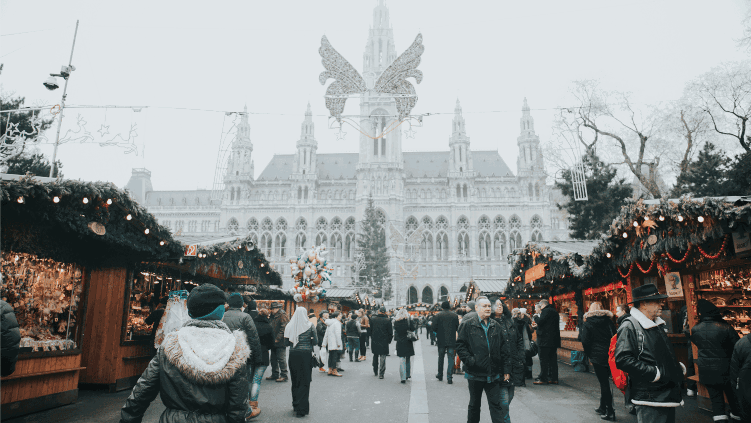 People stroll through a festive Christmas market with stalls on both sides, set against the backdrop of a grand, ornate building. A large decorative angel figure hangs overhead.