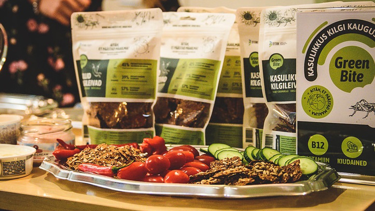 A table displays a silver tray with sliced cucumbers, cherry tomatoes, and crackers, alongside several bags of Green Bite brand products. The bags feature green and white labeling. A blurred person stands in the background.