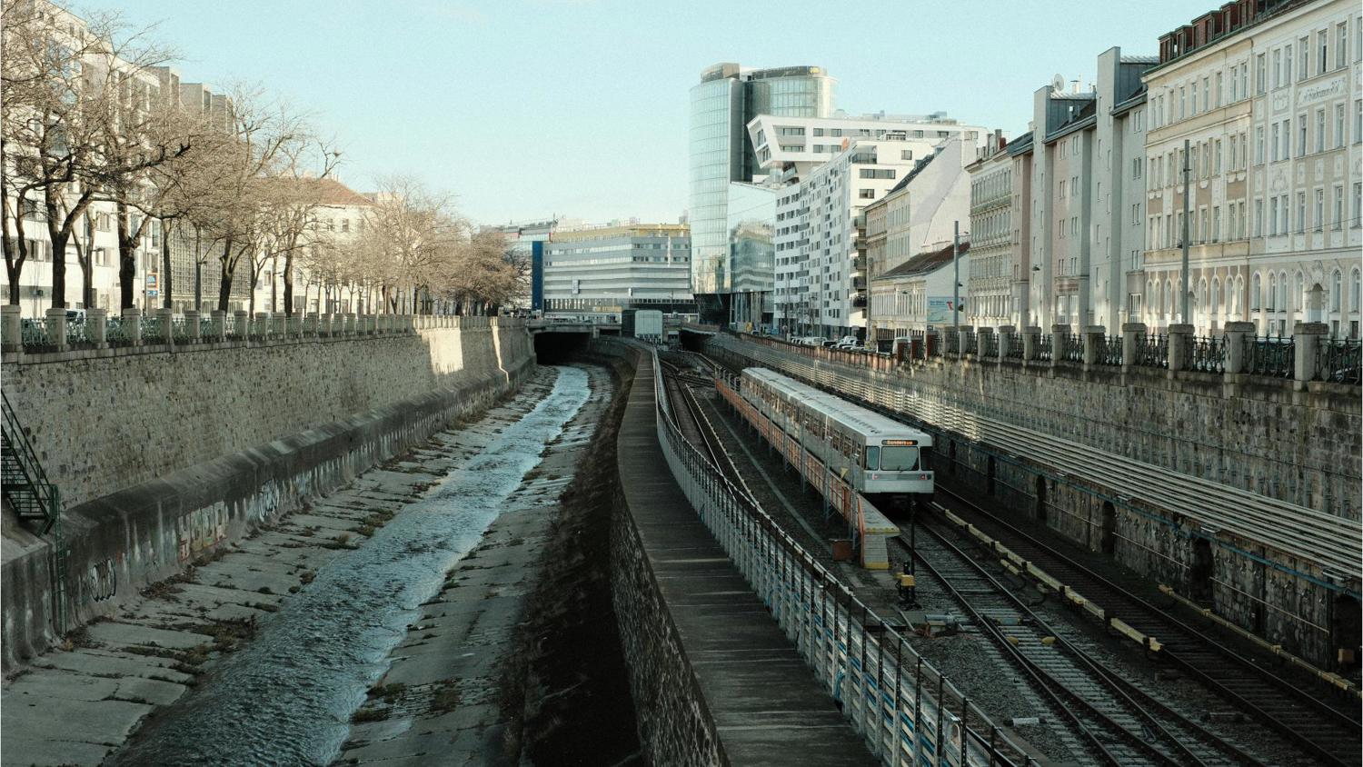 Train tracks run alongside a narrow water channel in an urban area. Tall buildings line both sides, with trees on the left. A train is on the tracks, and the sky is clear and blue.