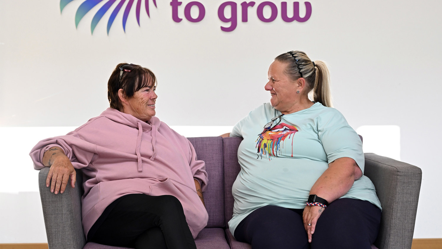 Two women sit on a gray couch, smiling at each other. Behind them is a wall with a colorful logo and the words "a space to grow." The setting appears bright and welcoming.