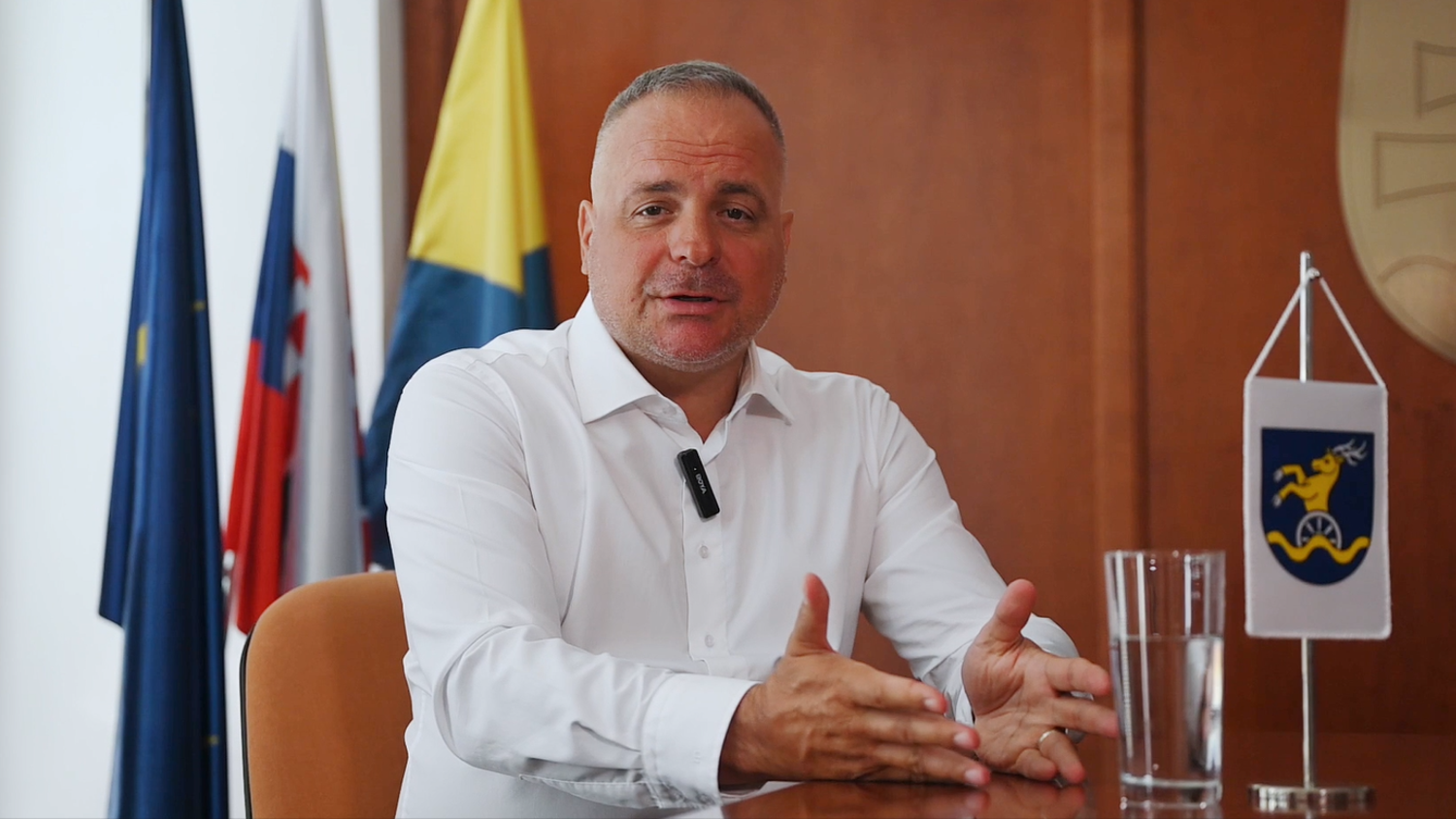 A man in a white shirt sits at a table, gesturing with his hands. There are flags and a glass of water on the table, with a microphone clipped to his shirt, in a formal office setting.