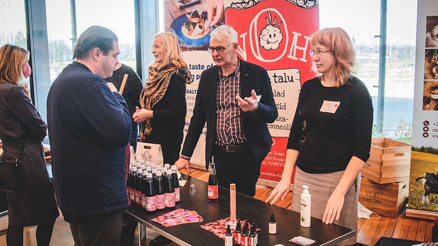 People are interacting at a booth displaying various bottled products and promotional materials. A banner with a logo is in the background. A man gestures while talking to others around a table. The setting appears to be an indoor event or exhibition.